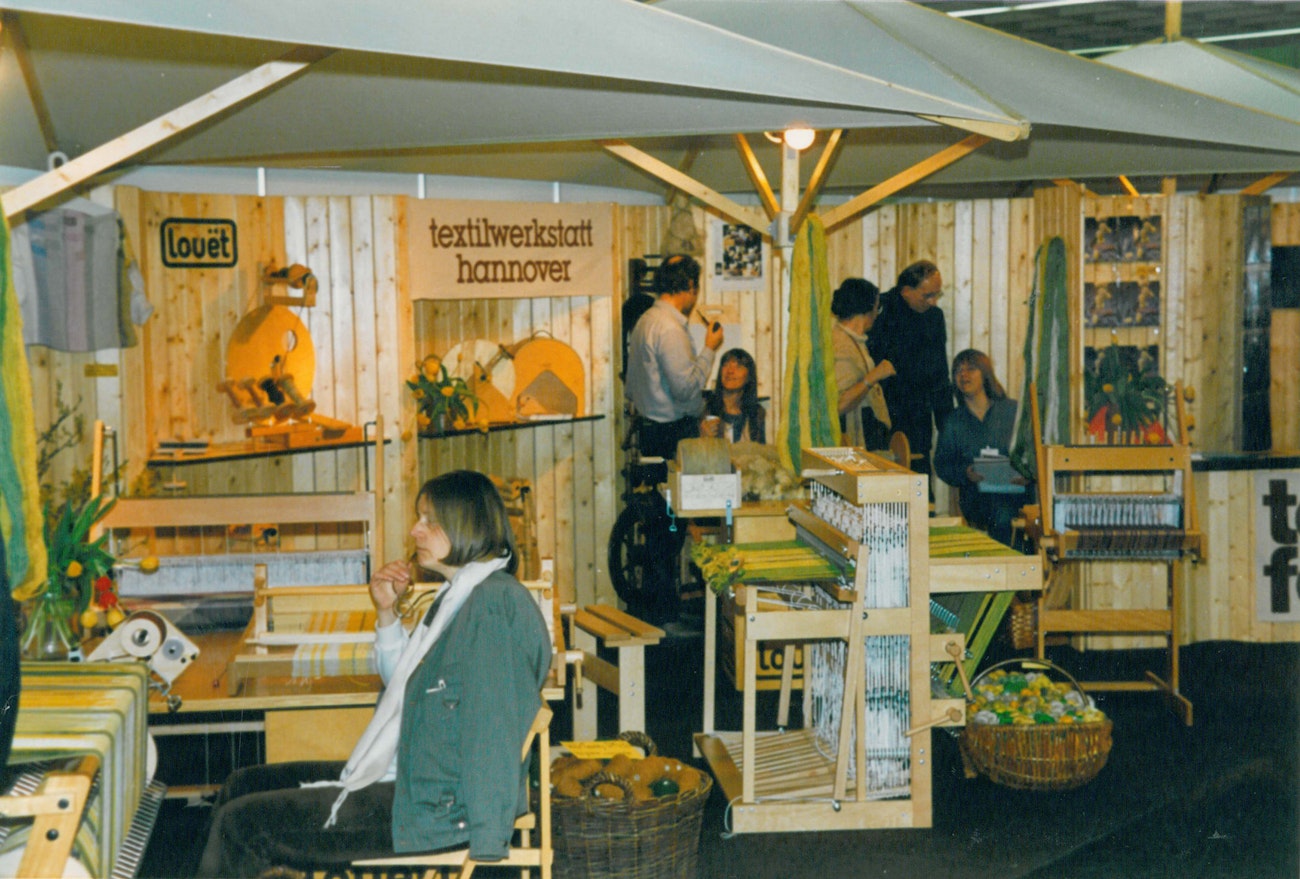 Louët wood-paneled showroom with spinning wheels on the wall, 5 looms on the floor, and a drumcarder bolted to a table in the background.
