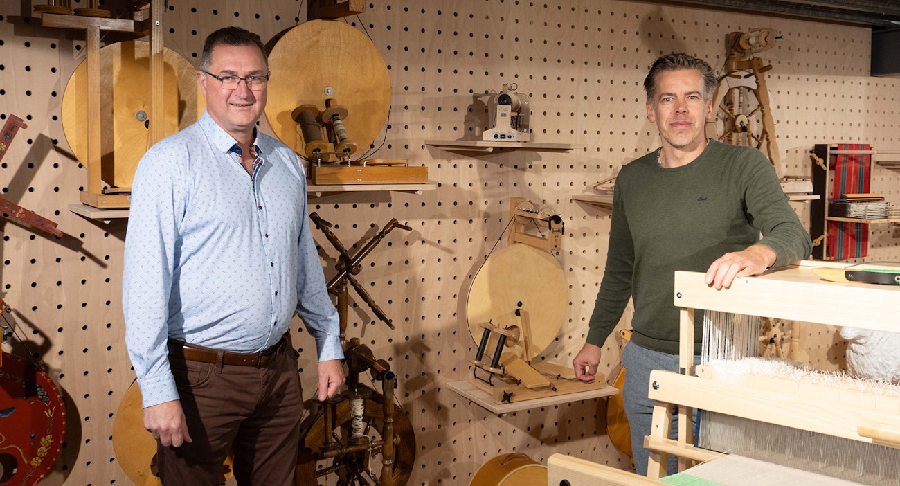 Two men standing before a wooden wall covered with spinning wheels, with a loom in the foreground.