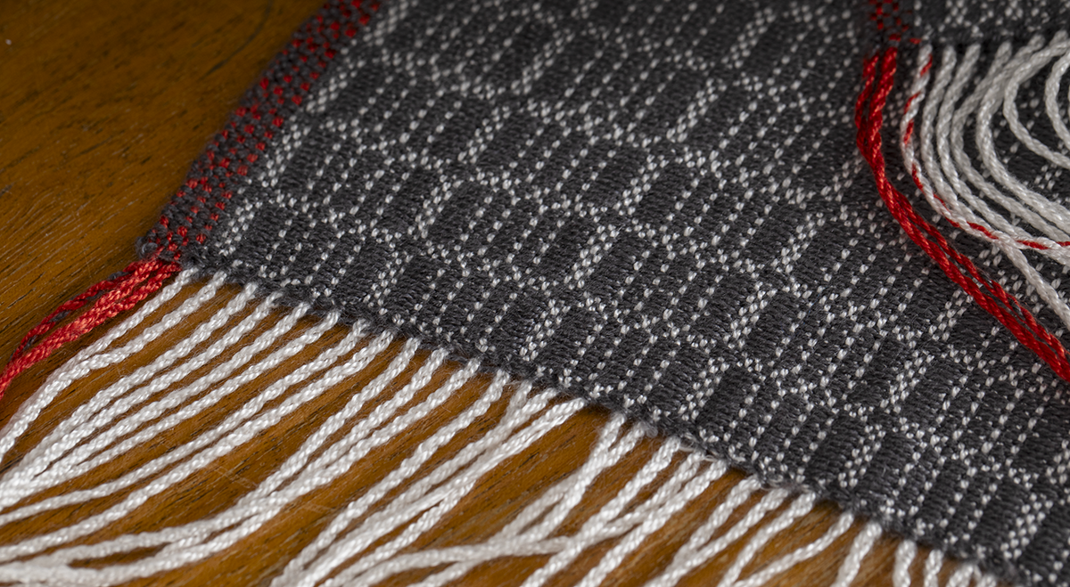 A scarf with grey and black patterning, and deep red edges draped on the side of a handbag.