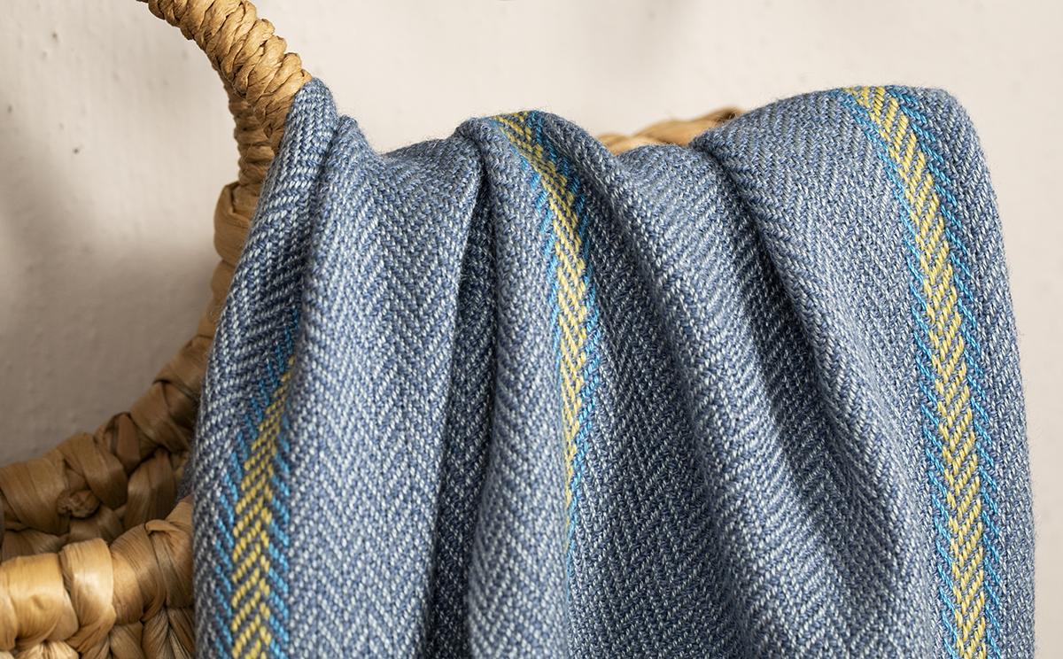 A scarf with blue, gray, and yellow herringbone patterning draped on the side of a basket.